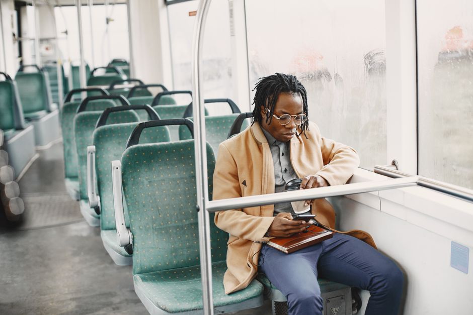 African American man on bus, wearing facemask and coat, using smartphone for commuting.