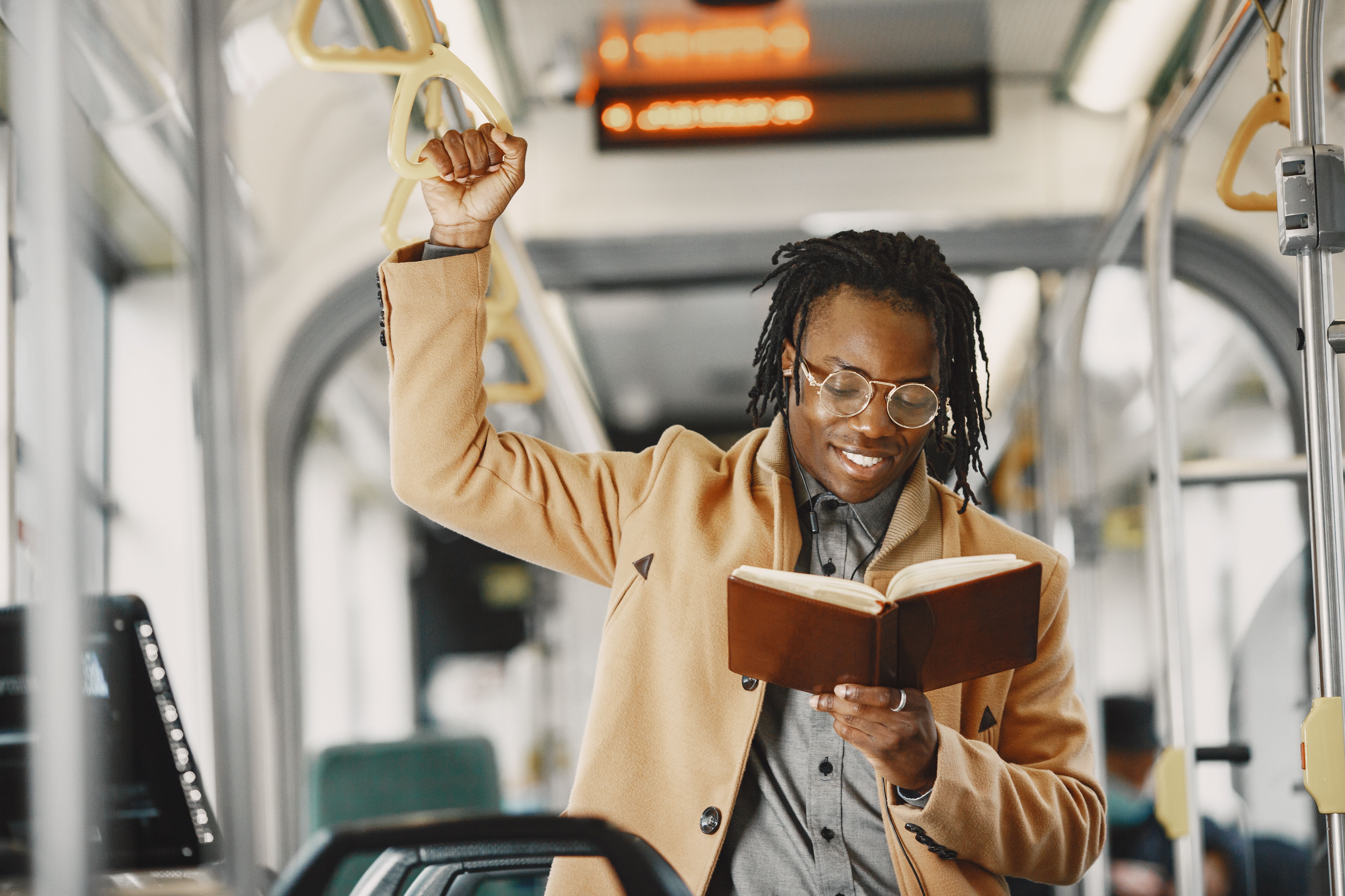 Smiling man with eyeglasses reading a book on an urban bus commute.