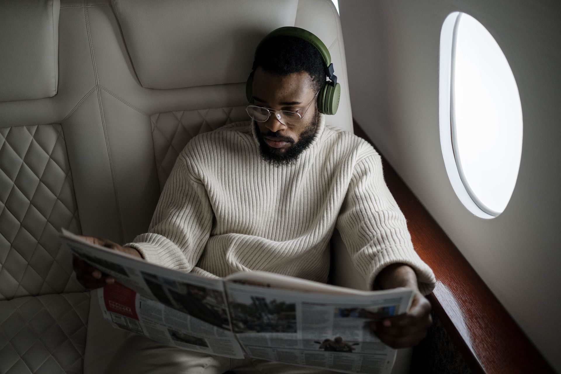 Business man reading newspaper on airplane wearing headphones and glasses.