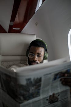 An African American businessman wearing headphones reads a newspaper on a flight.