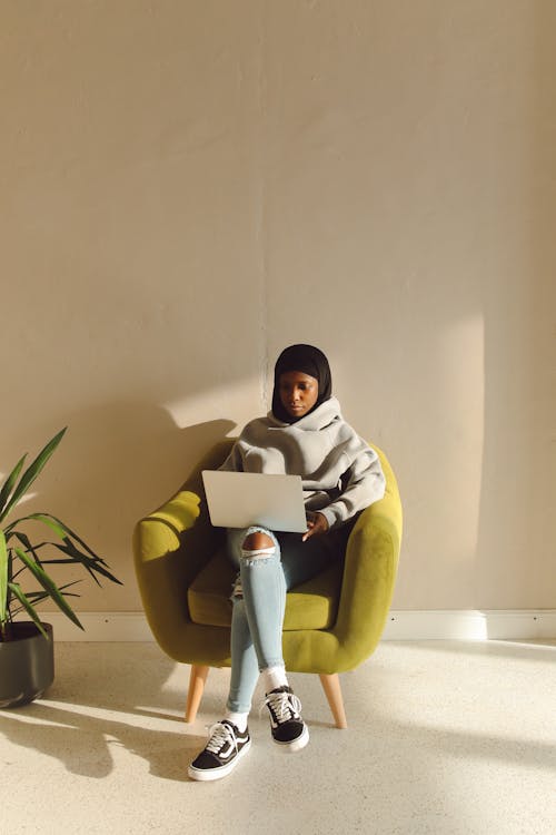 Free A Woman Wearing a Hoodie Jacket Sitting on the Couch while Using Her Laptop Stock Photo