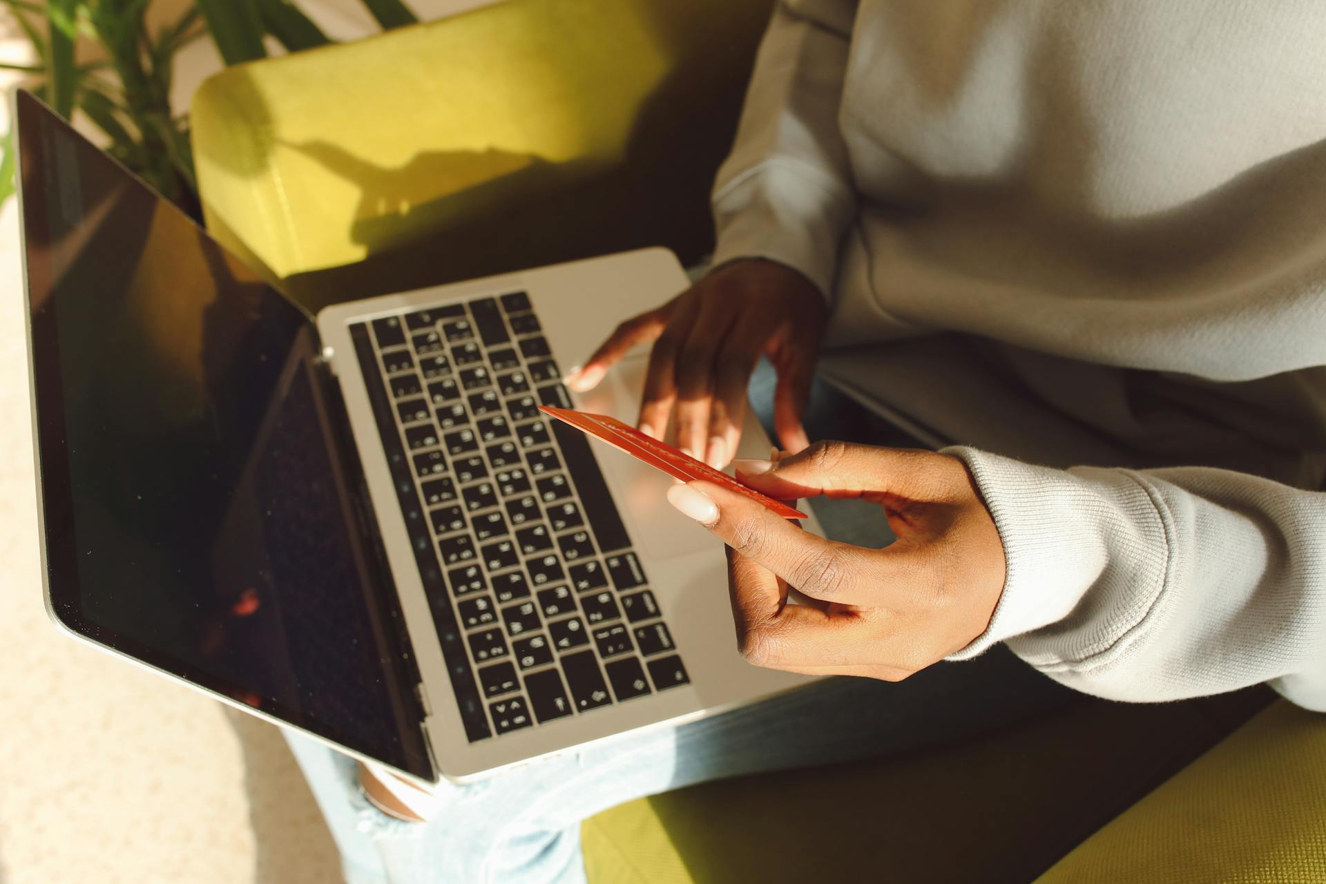 Person using credit card for online shopping on a laptop while seated indoors, sunlight streaming in.