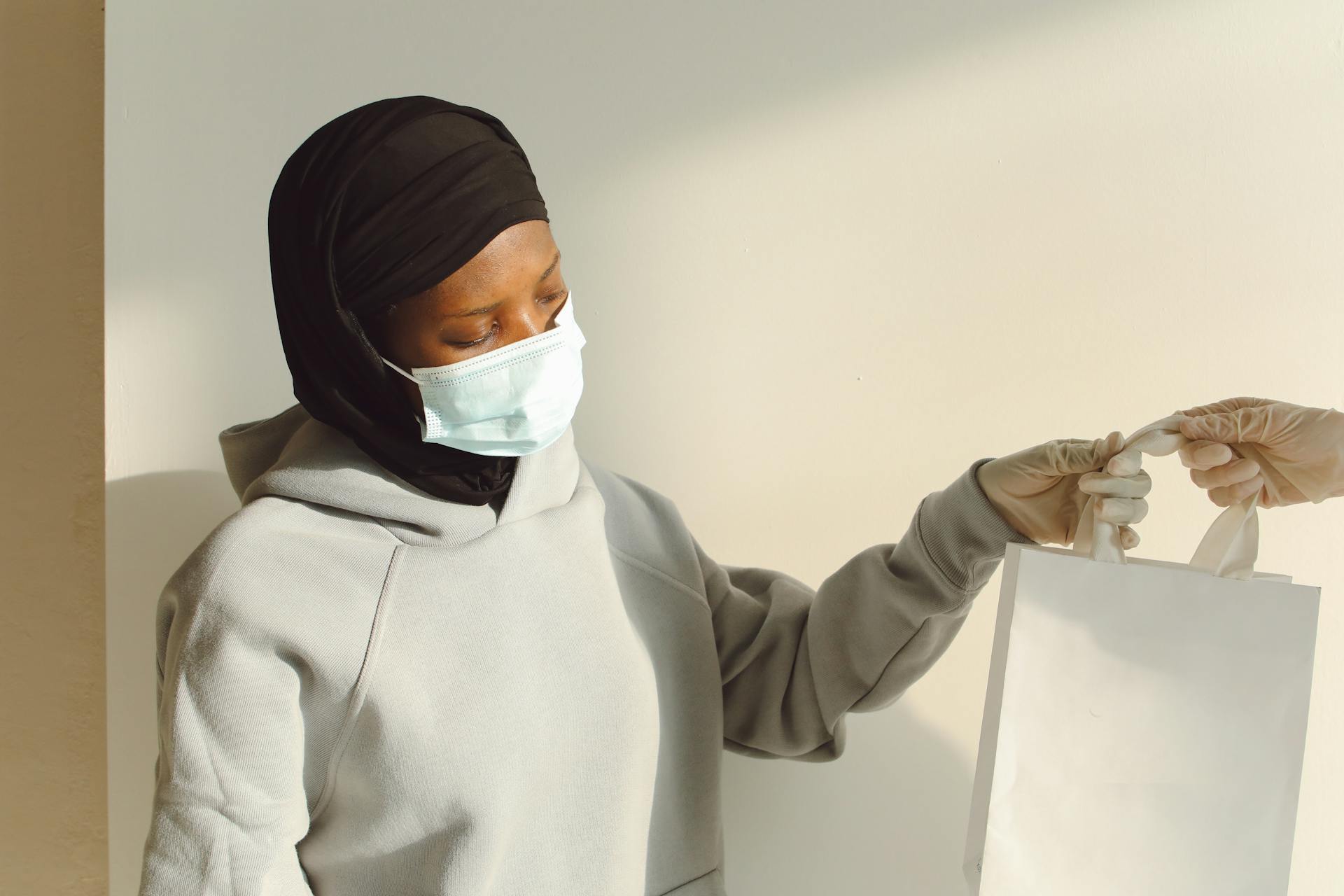 A woman in a hijab and mask receiving a delivery bag from a gloved hand.