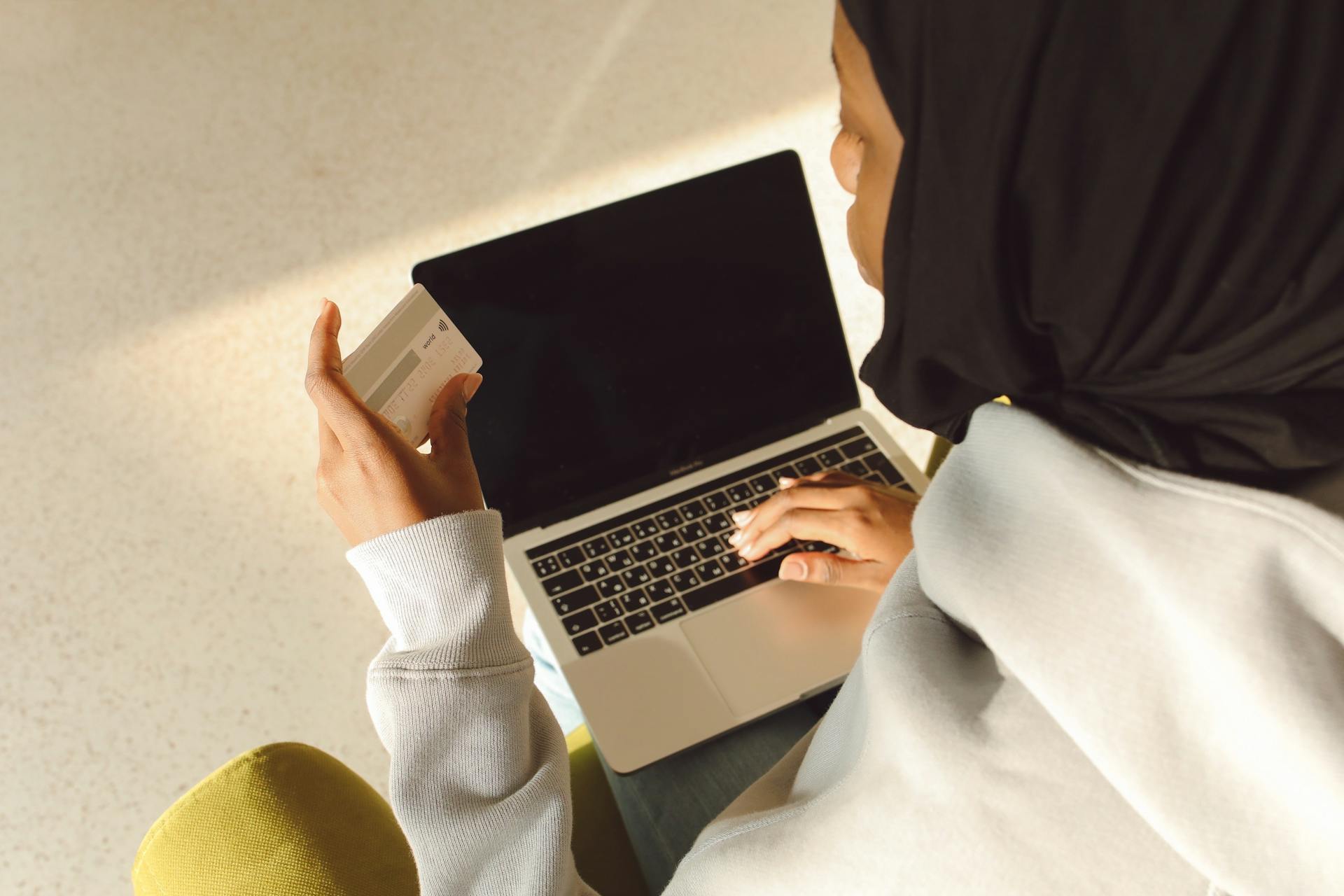 A woman in a hijab using a laptop and credit card indoors for online shopping.