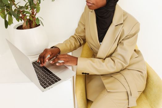 Stylish woman in beige suit typing on laptop indoors, near a plant.