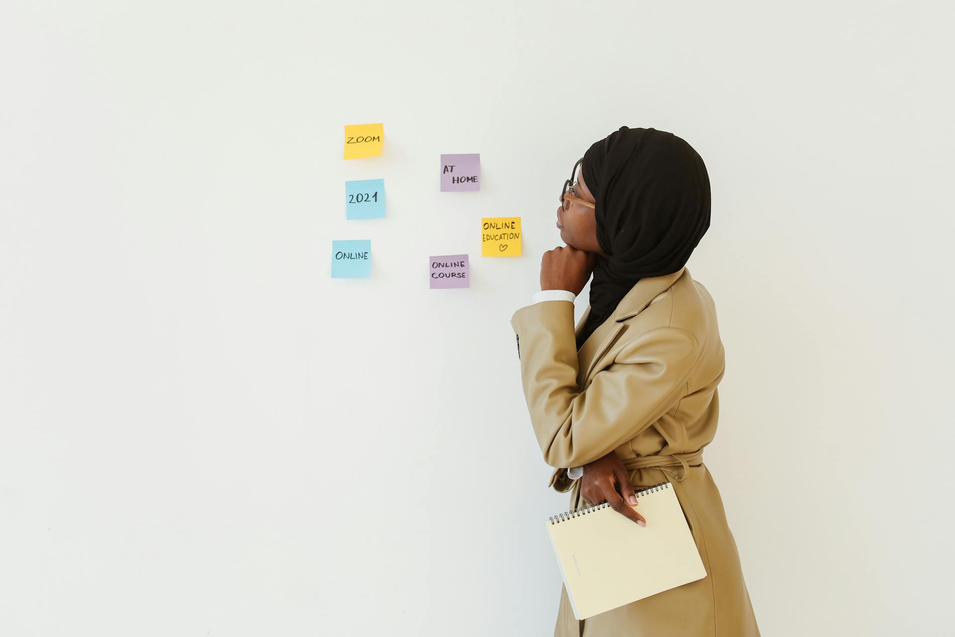 A woman in a hijab stands indoors, contemplating sticky notes about online courses and education.