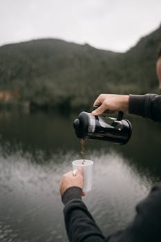 A person pours coffee from a thermos beside a calm lake, enjoying a serene outdoor moment.