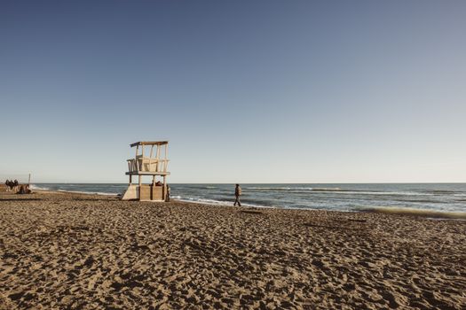 A serene beach setting with a lifeguard hut and people strolling under a clear blue sky.