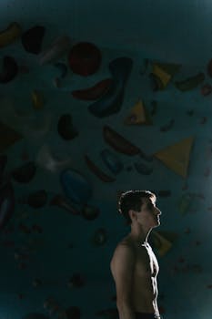 Side profile of a shirtless man in an indoor rock climbing gym with colored holds.