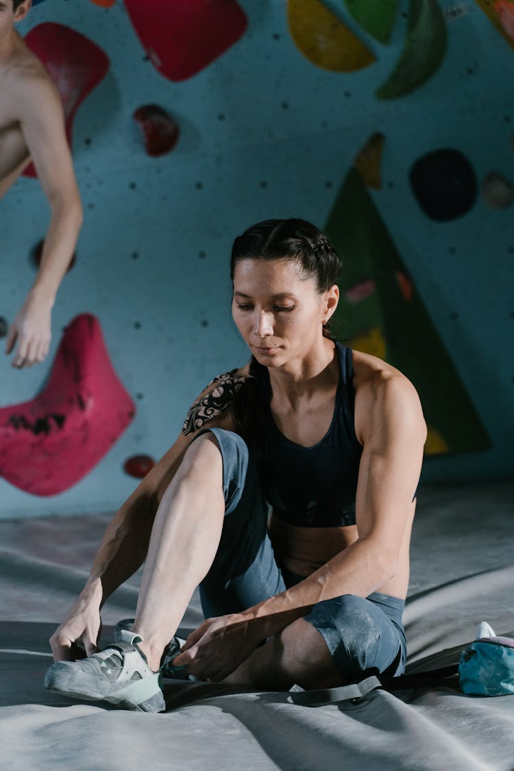 Woman Putting On Climbing Shoes Sitting On A Mattress By The Climbing Wall