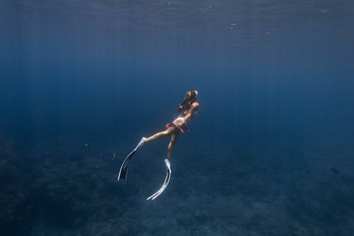 Side view full body of anonymous sporty female diver in flippers and mask swimming underwater of blue sea near coral reefs