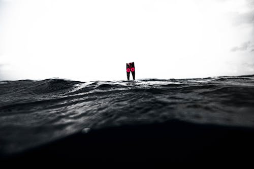 Black and white of unrecognizable person in flippers swimming underwater of wavy ocean against cloudy sky