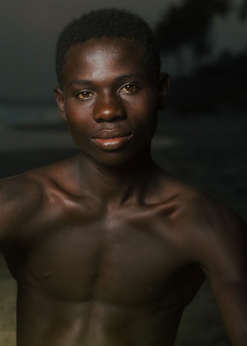 Self assured muscular African American male millennial with naked torso looking at camera while standing on sandy beach in dark evening