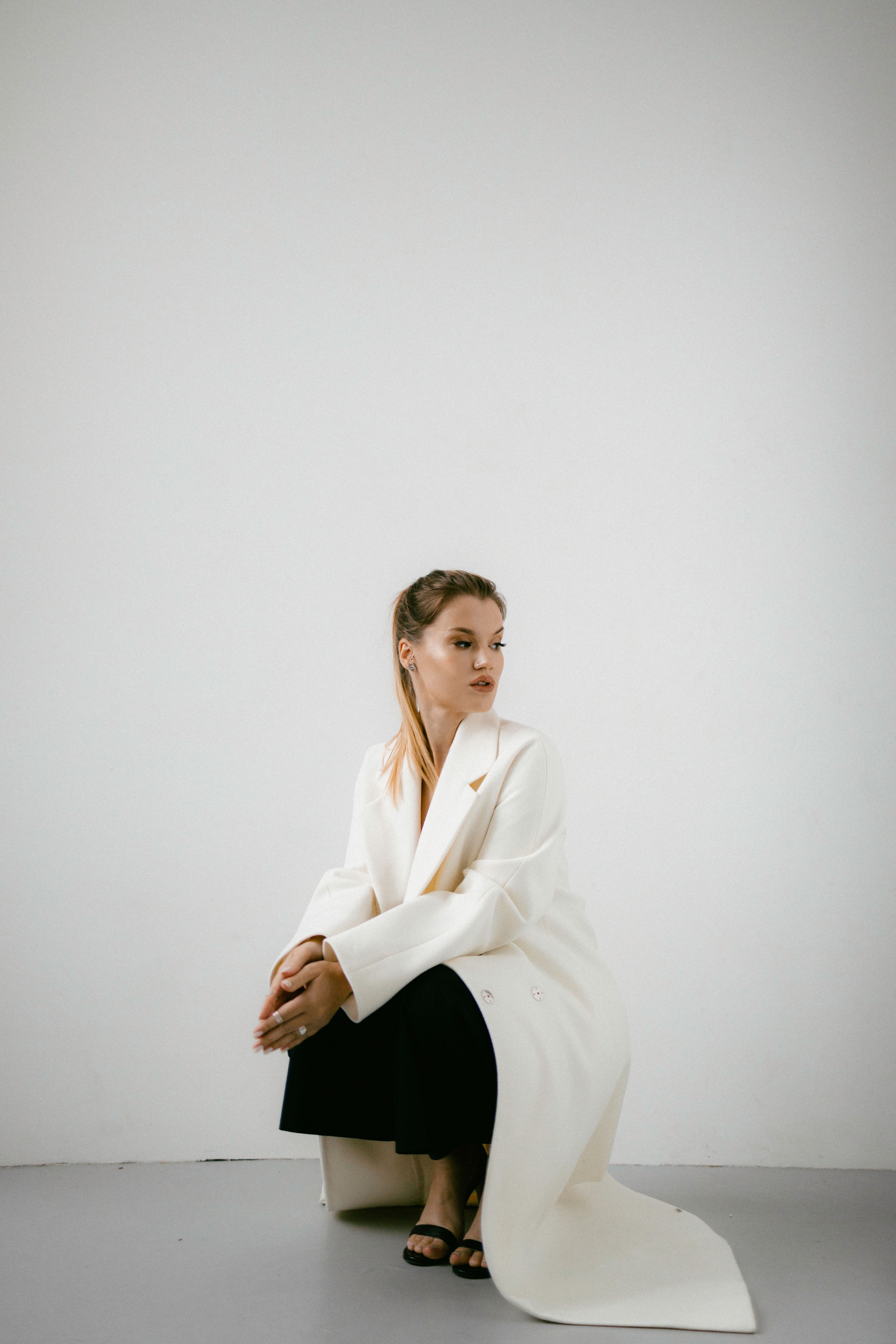 Stylish woman in a white coat squatting against a minimalist white wall indoors.