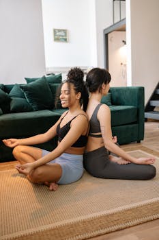 Two women meditating in lotus pose on a rug indoors, back to back with eyes closed.