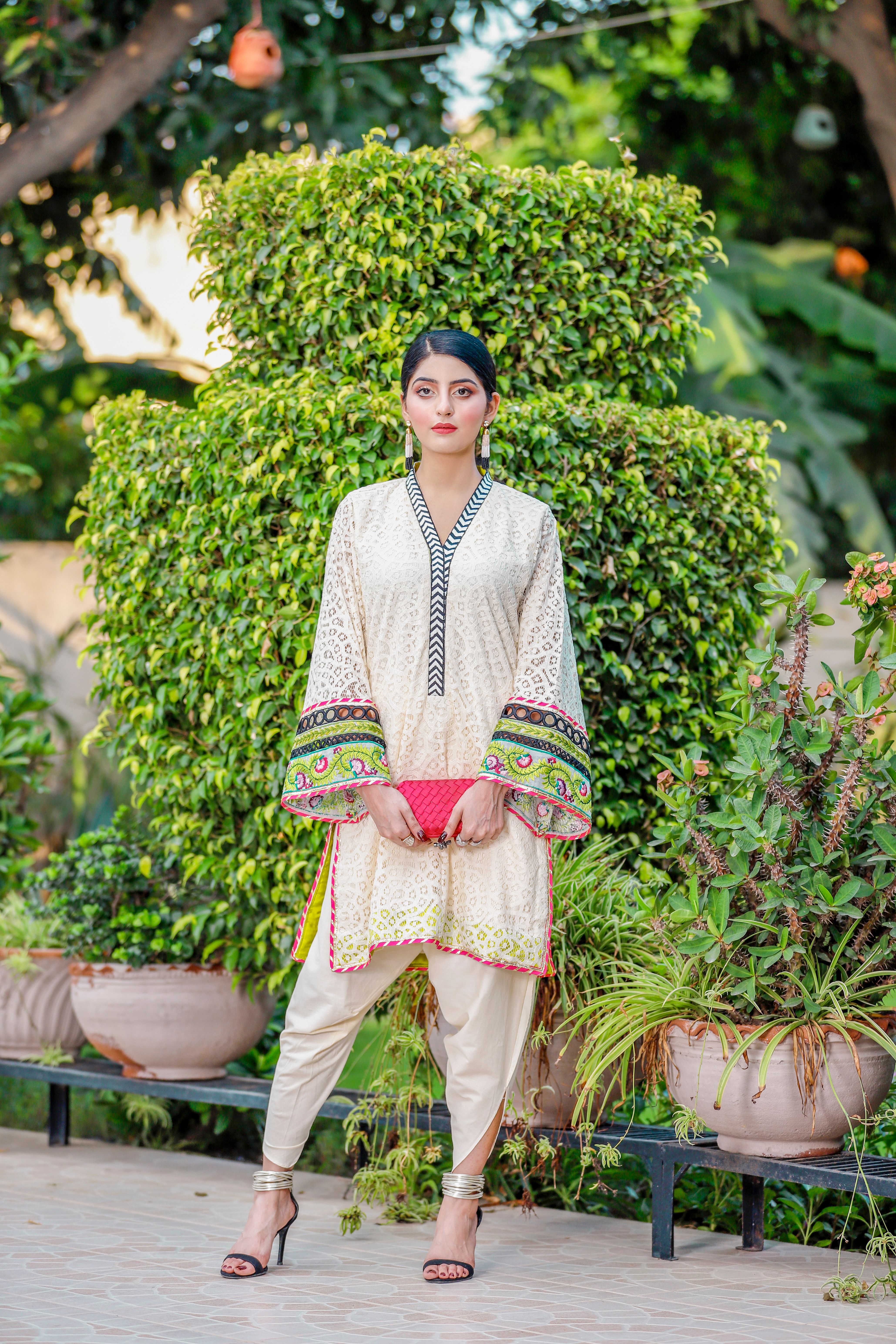 Asian woman in a traditional outfit stands in a garden with lush greenery.
