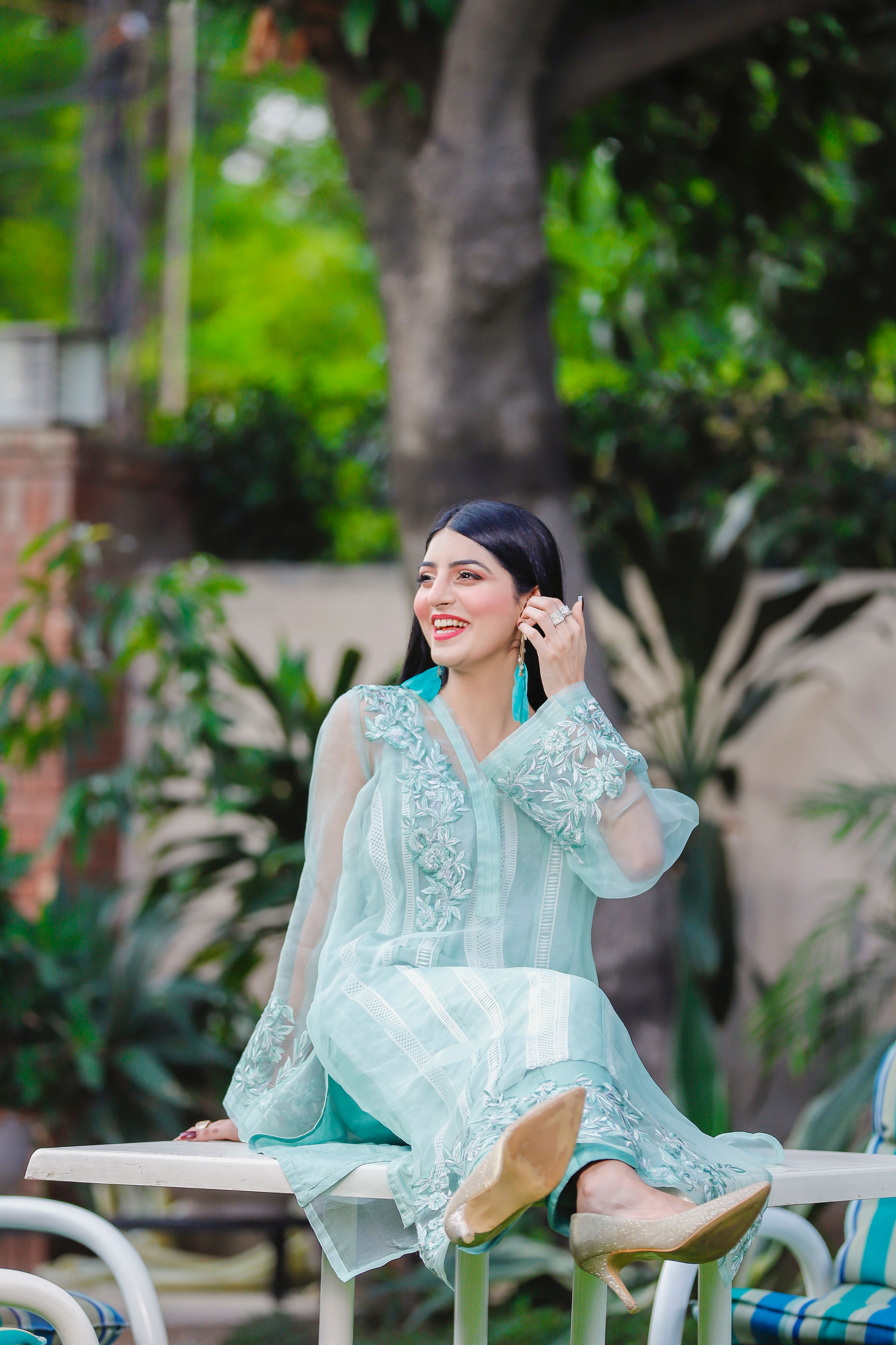 A cheerful woman in a traditional dress sitting outdoors, surrounded by lush greenery.
