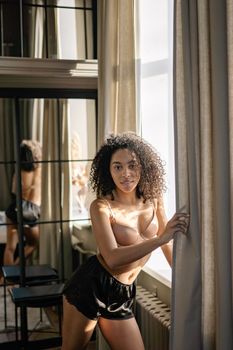 A beautiful woman with curly hair posing confidently by a window, creating a dramatic indoor scene.