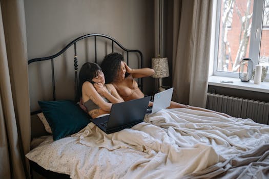 Two women working on laptops from home in a cozy bedroom setting, embodying remote work lifestyle.