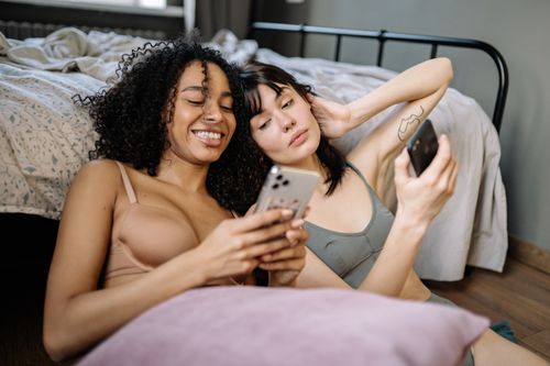 Free Two women relaxing at home, sharing smiles and phones, creating joyful moments together. Stock Photo