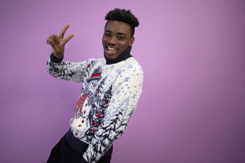 Free Smiling man in a winter-themed sweater against a purple background making a peace sign. Stock Photo