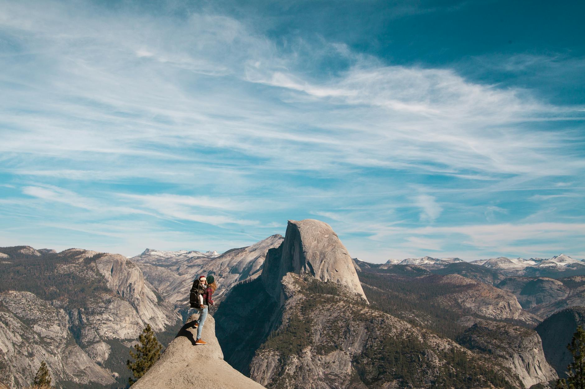 A woman and child stand atop Half Dome, Yosemite, with breathtaking views of the mountain range.