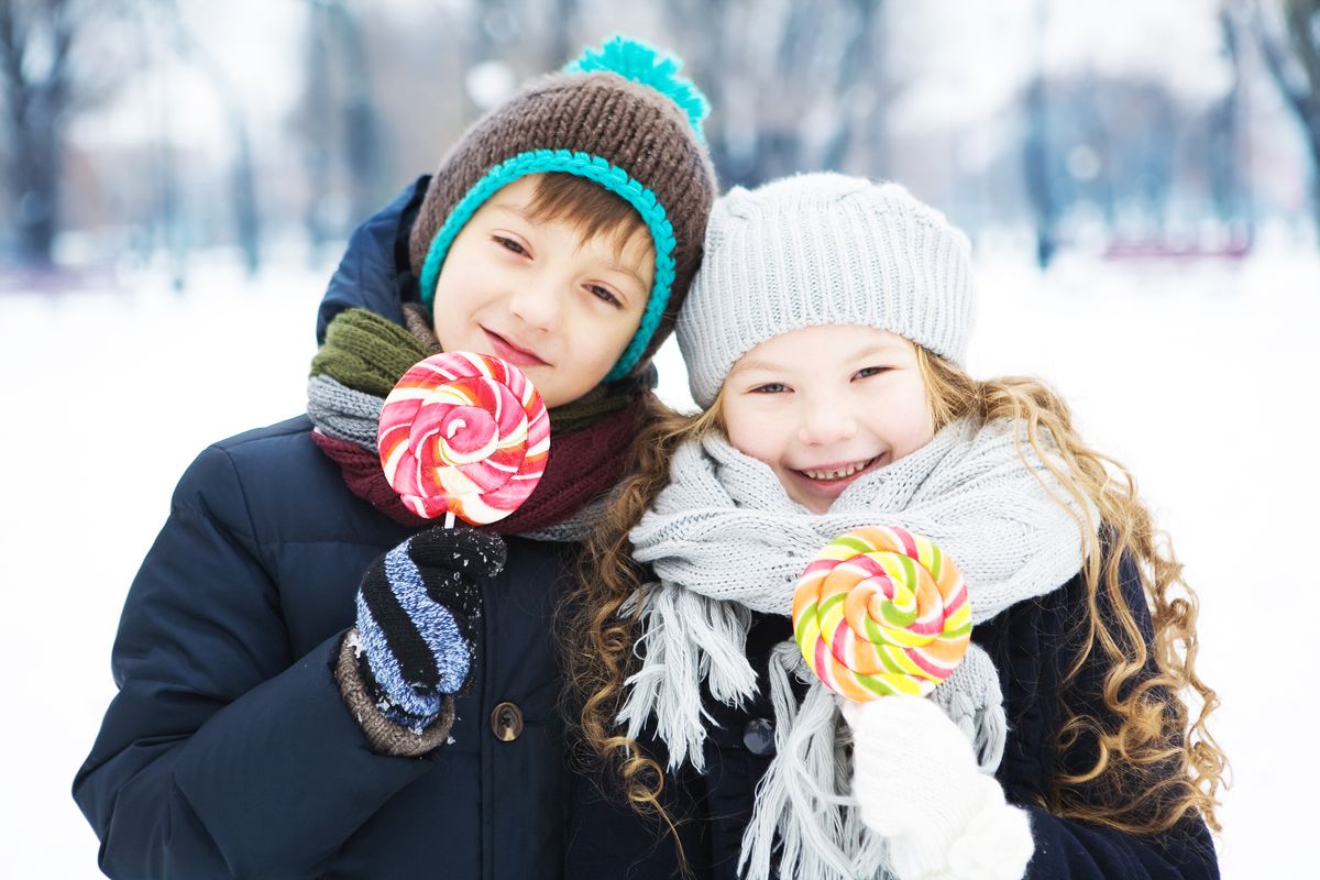 Two children happily enjoy lollipops in winter wear, smiling in a snowy outdoor setting.