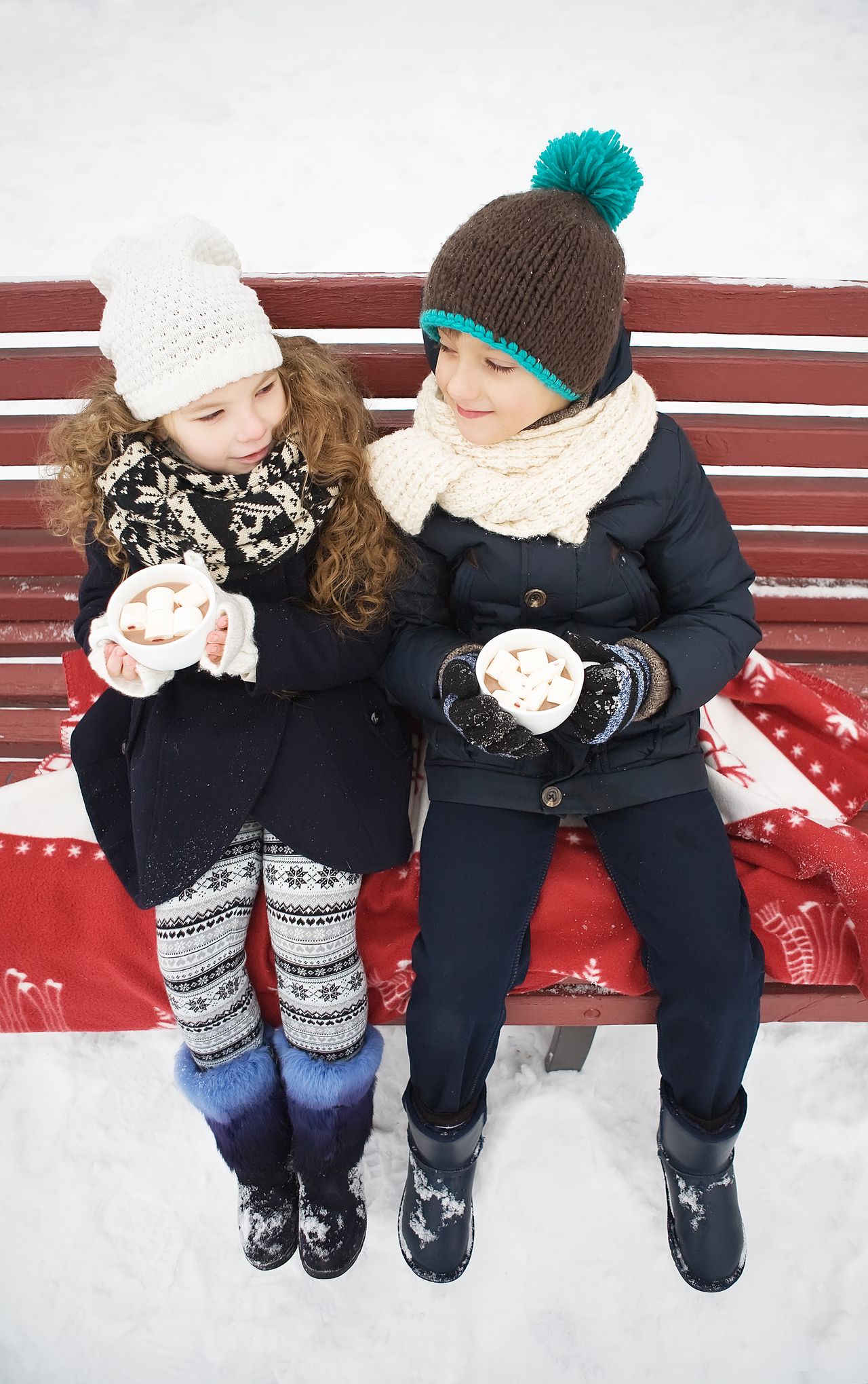 Two children in winter boots sitting on a snowy bench drinking hot cocoa