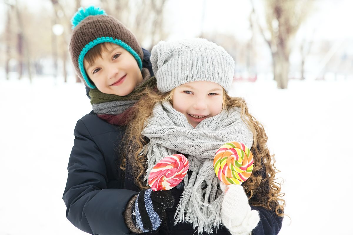 Smiling siblings in winter attire hold colorful lollipops outdoors in snowy scenery.