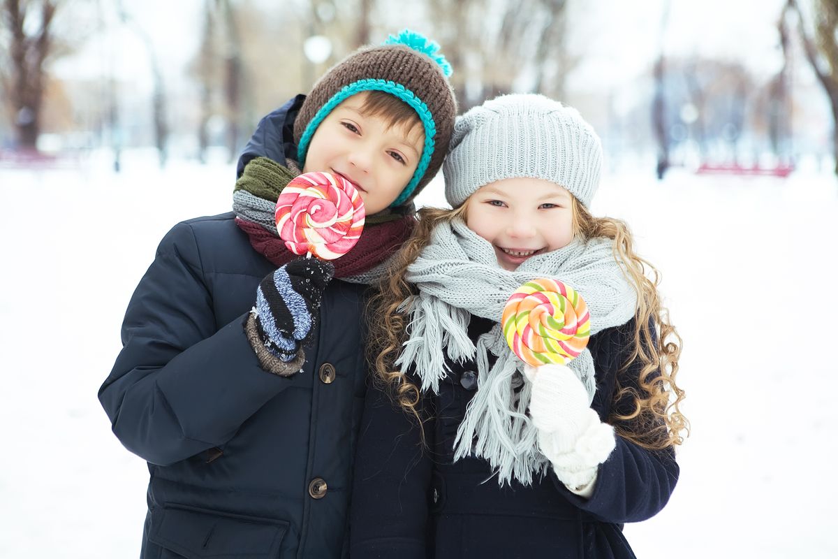 Smiling children wearing winter clothes enjoy lollipops outside in a snowy park.
