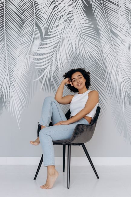 Smiling woman with curly hair in casual attire sitting on a chair against a leaf-patterned background.