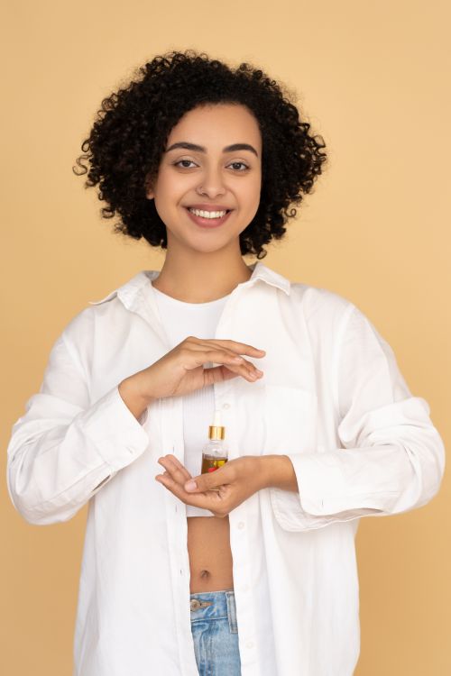 Free Smiling woman with curly hair holding a bottle of essential oil indoors, promoting wellness and beauty care. Stock Photo