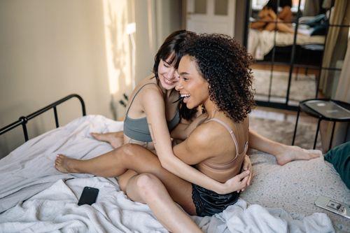Free Happy lesbian couple sitting and laughing on bed, embracing love and joy indoors. Stock Photo
