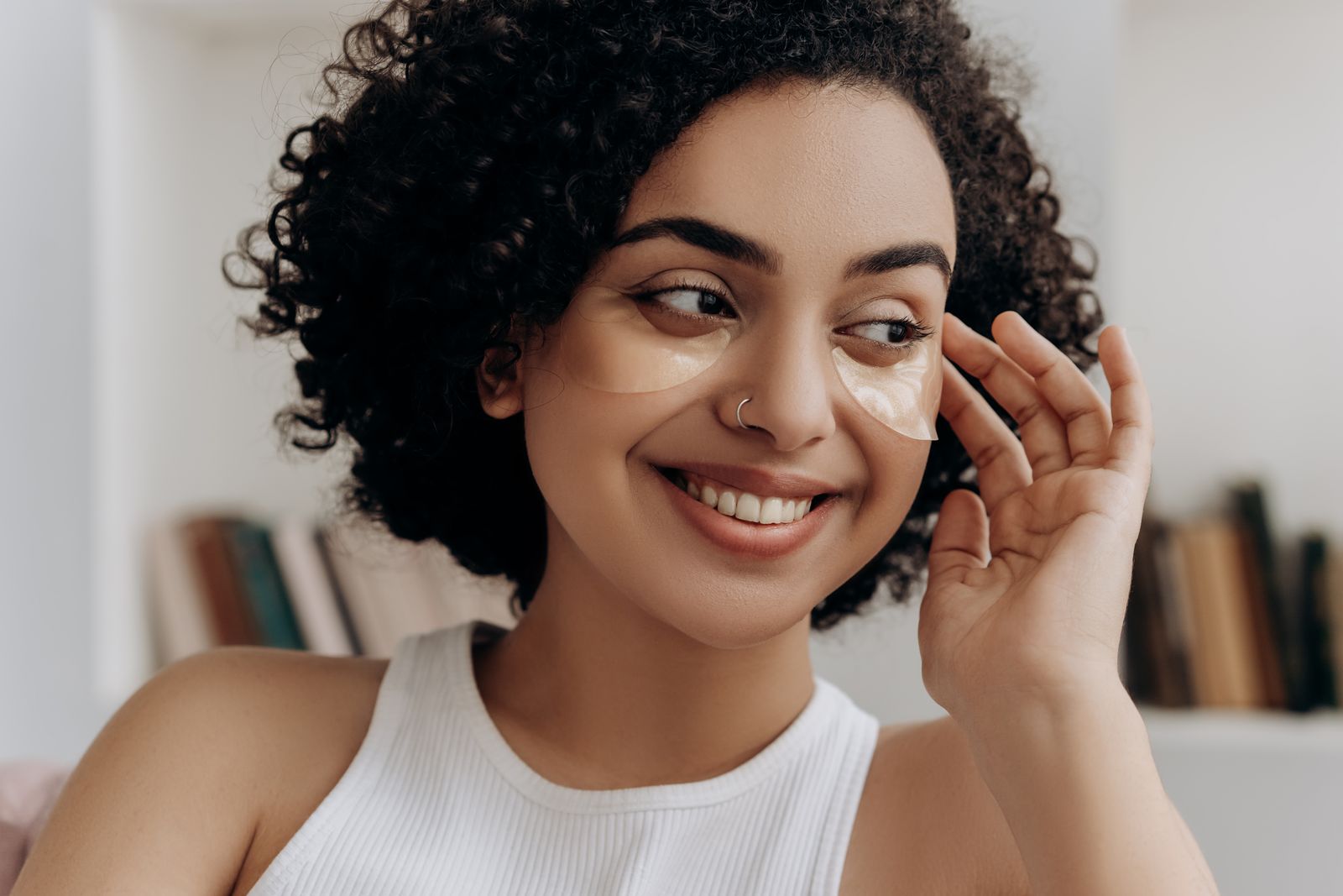 Smiling woman with curly hair wearing clear under-eye patches, touching her cheek in a bright room
