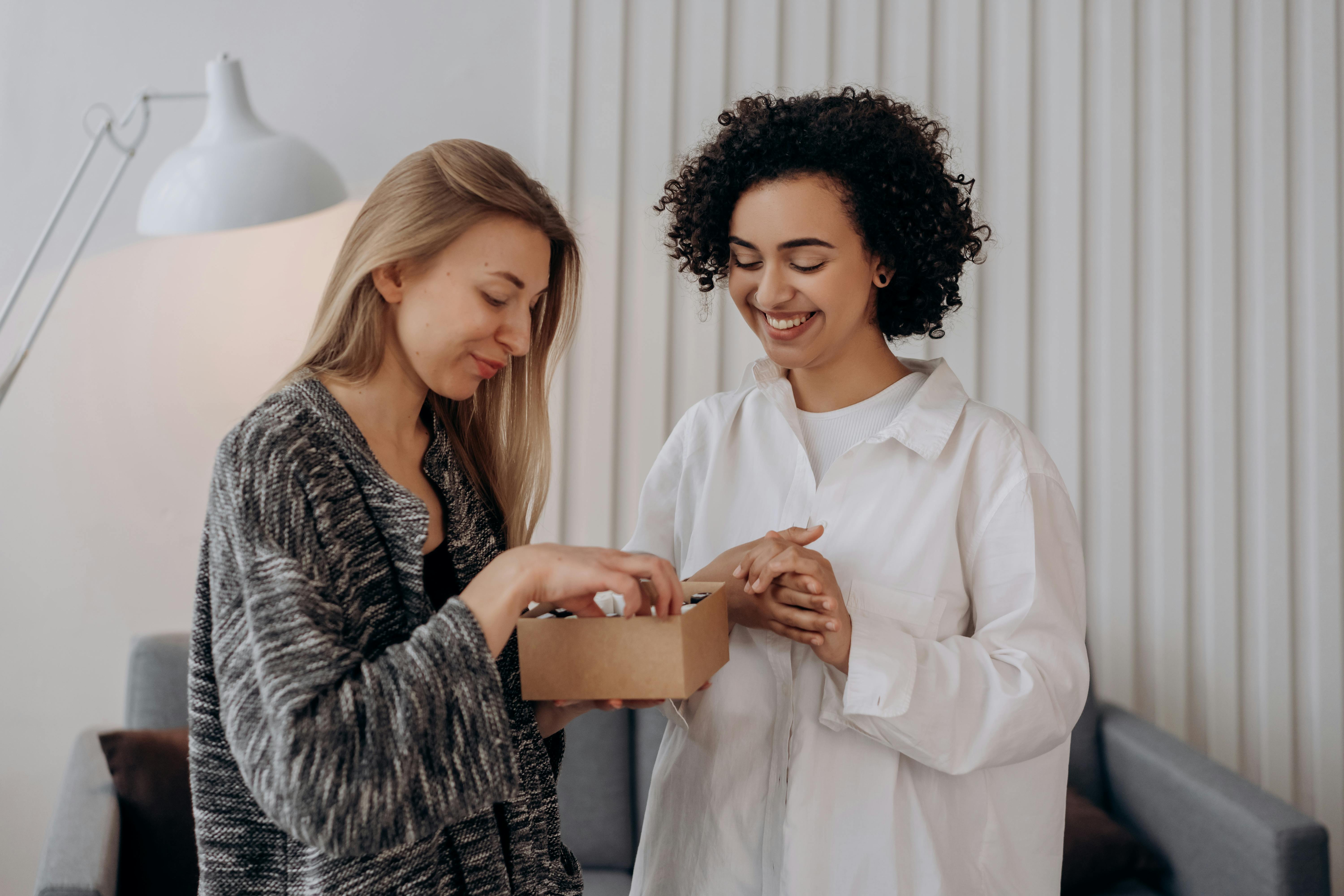 Two women smiling while exchanging a gift box indoors. Perfect for lifestyle and wellness themes.