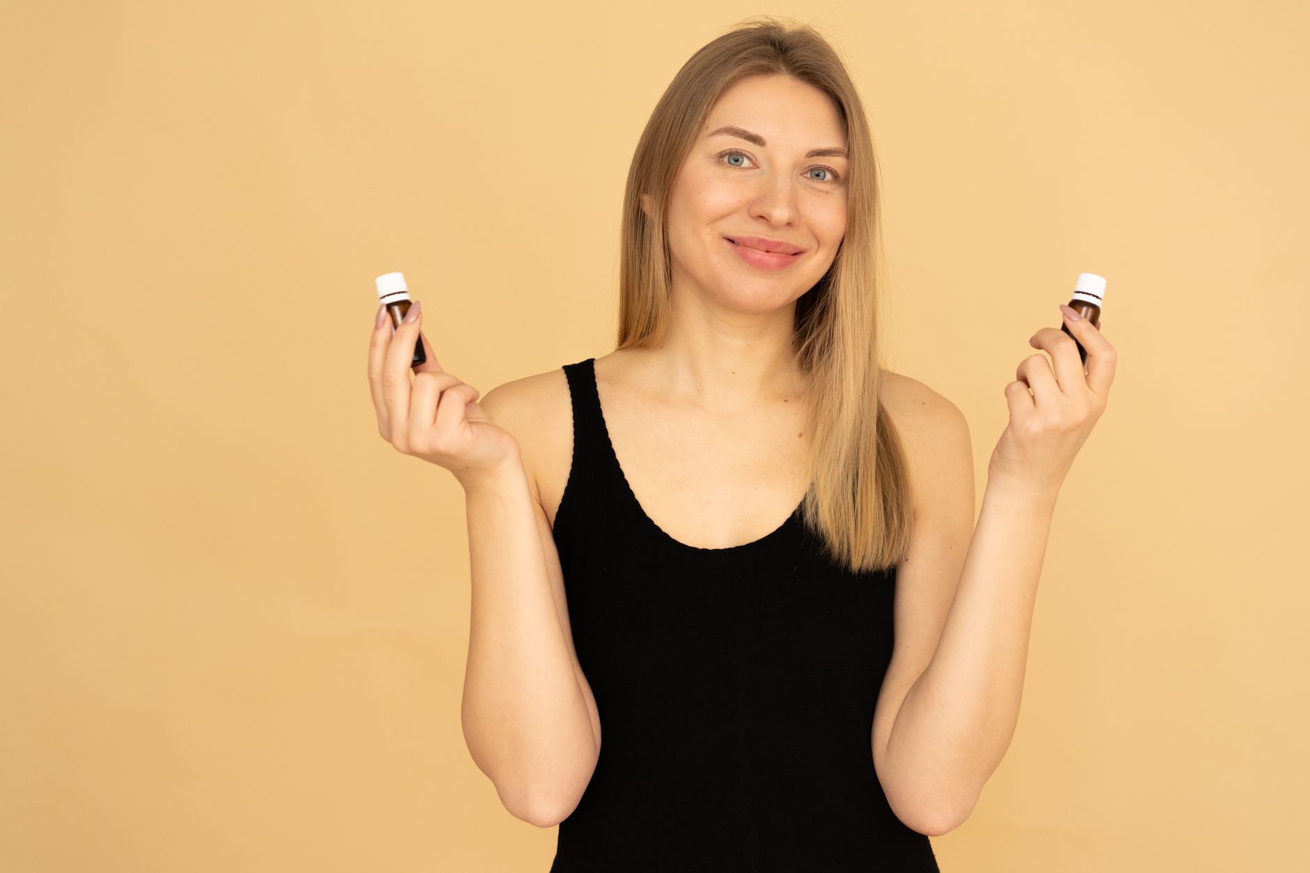 Woman holding essential oils with a joyful expression against a beige background, emphasizing wellness and beauty.