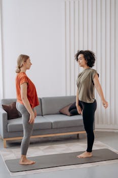 Two women standing on yoga mats, practicing yoga poses indoors, promoting friendship and mindfulness.