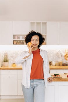 A woman stands in a modern kitchen holding an orange, promoting healthy lifestyle and cooking.