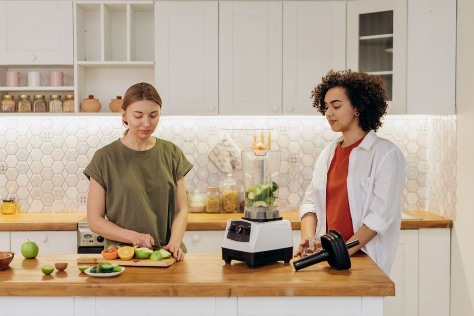 Two women preparing a healthy meal together in a modern kitchen, using fresh vegetables and a blender.