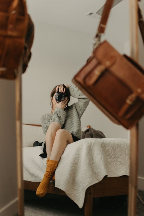 Free Unrecognizable young female sitting on bed while taking self shot in reflection of mirror in bedroom Stock Photo