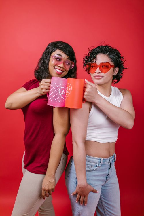 Free Smiling women in heart sunglasses toasting mugs against a red background, celebrating love and friendship. Stock Photo