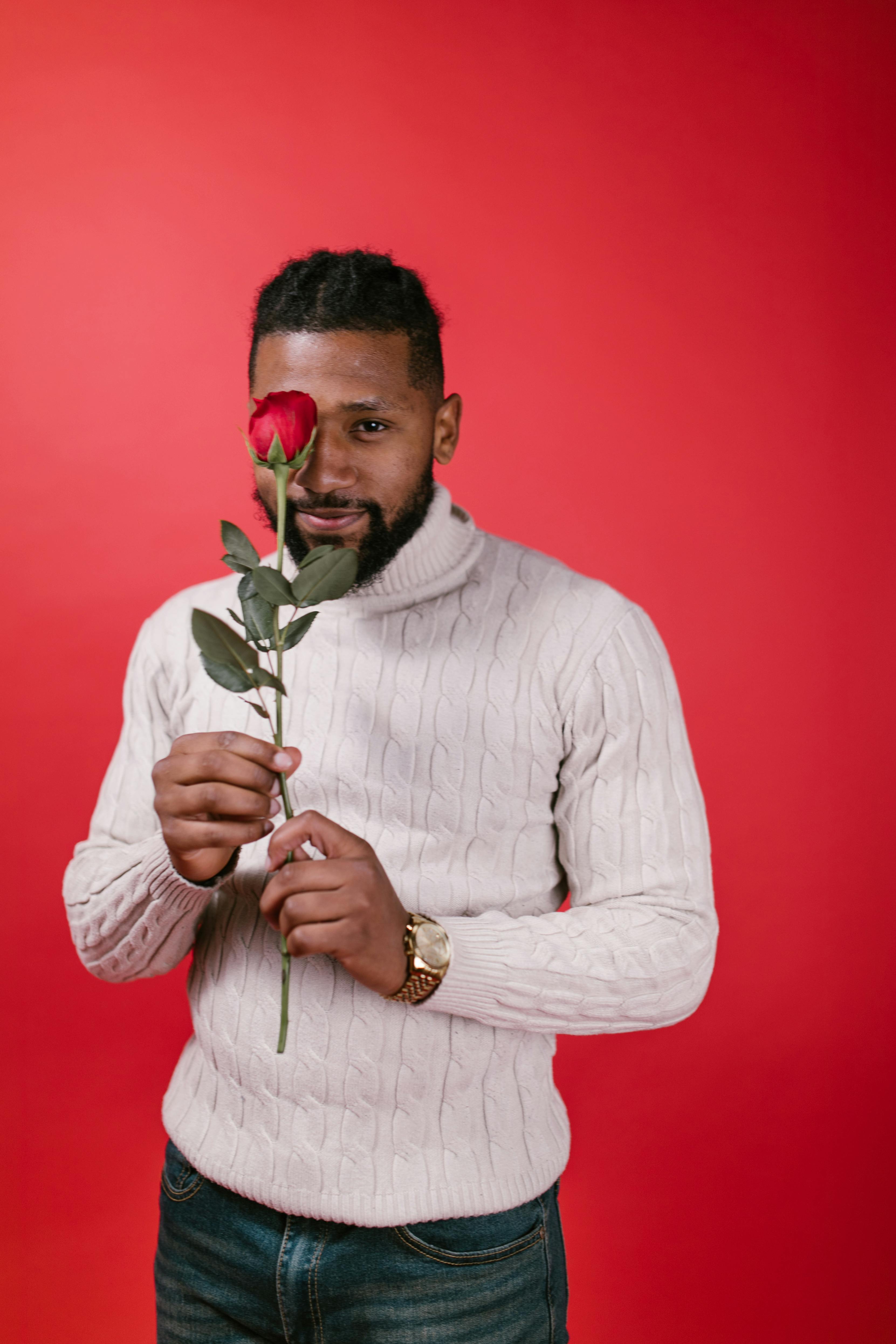 Man in white sweater holding red rose on a vibrant red background, perfect for Valentine's themes.