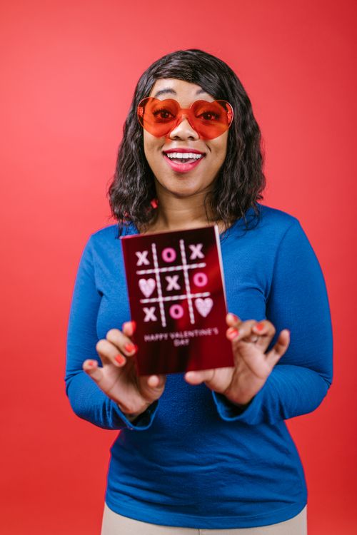 Free Smiling woman with heart sunglasses holding a Valentine's card against a red background. Stock Photo