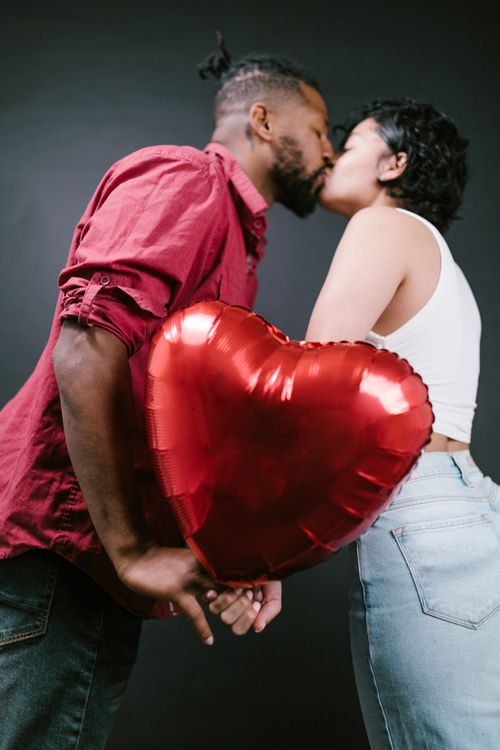 Free A romantic couple sharing a kiss while holding a red heart-shaped balloon. Stock Photo
