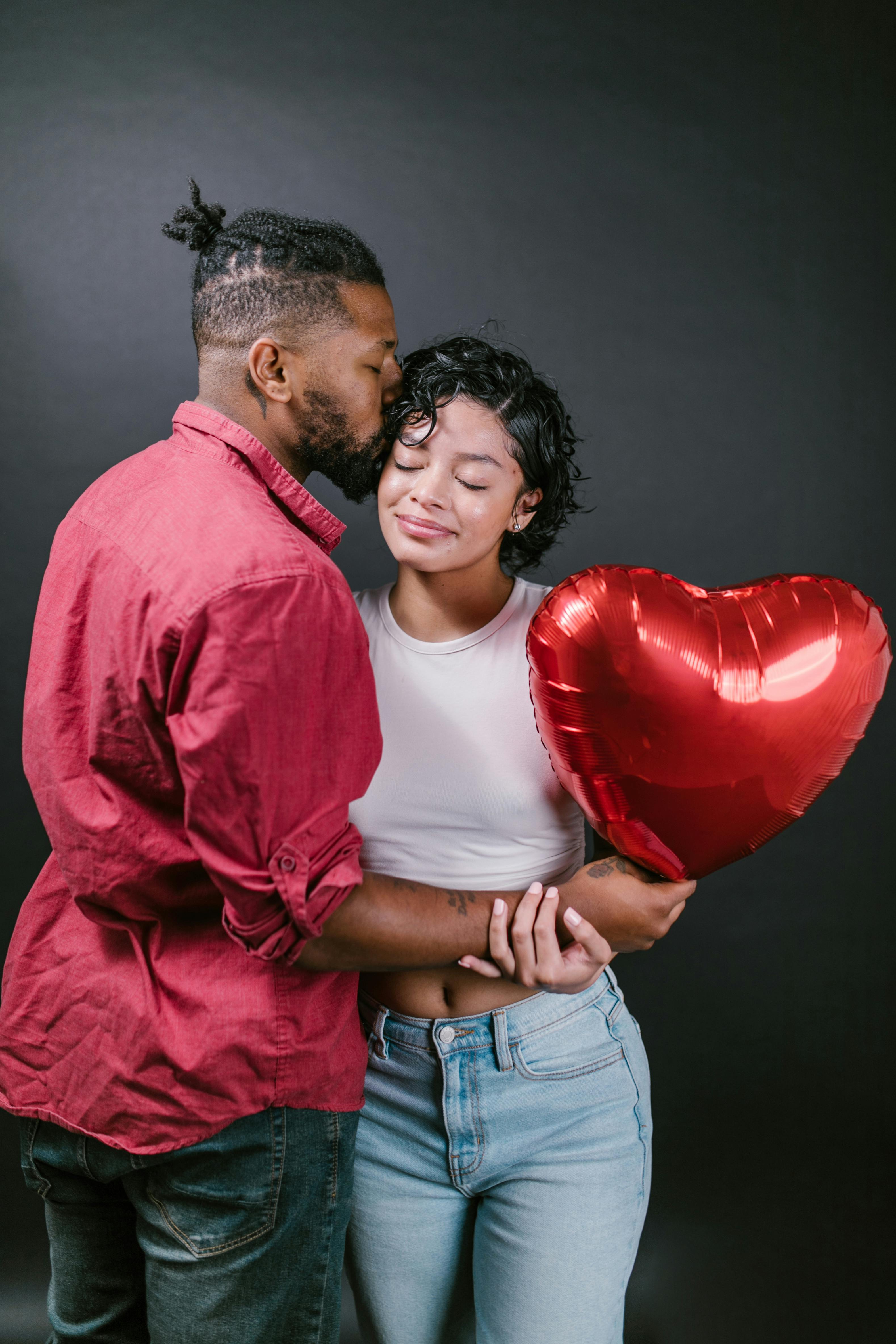 man kissing his woman while holding a red heart shaped balloon