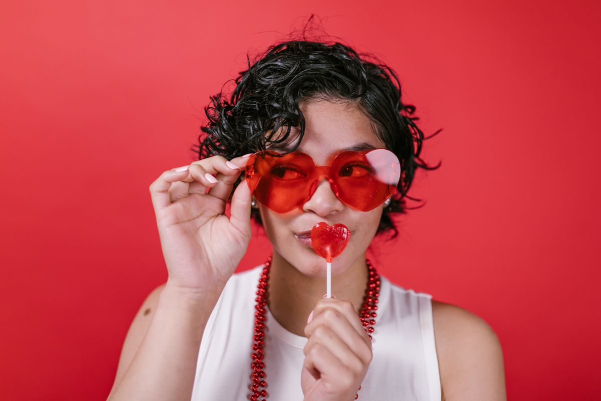 Portrait of a woman wearing heart shaped sunglasses and holding a lollipop on a red background.