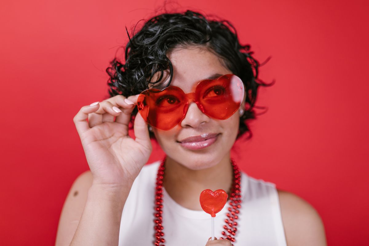 Woman in red sunglasses and necklace smiles while holding a heart-shaped lollipop against a red background.