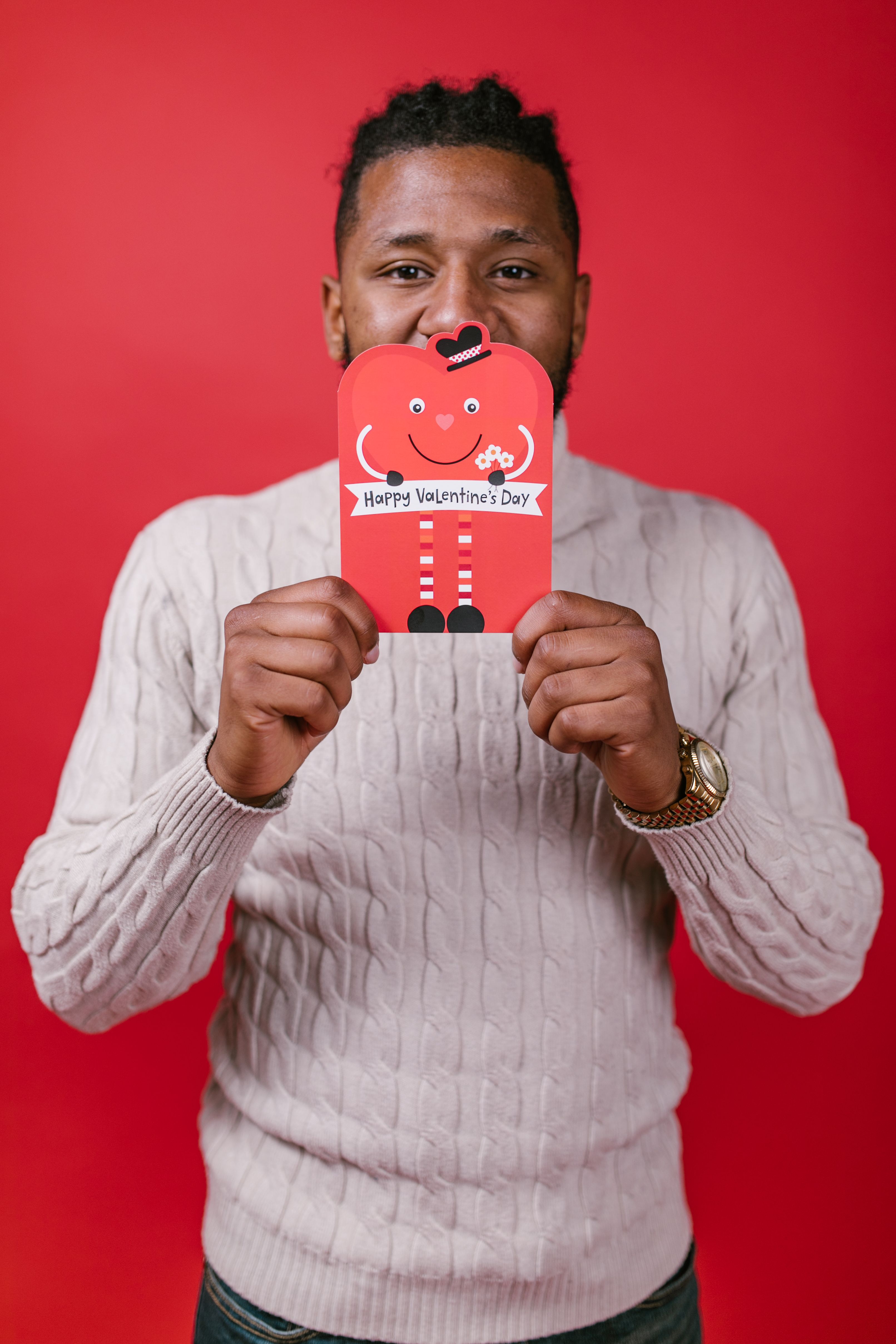 Man in sweater smiling and holding a Valentine's Day card against a red background.