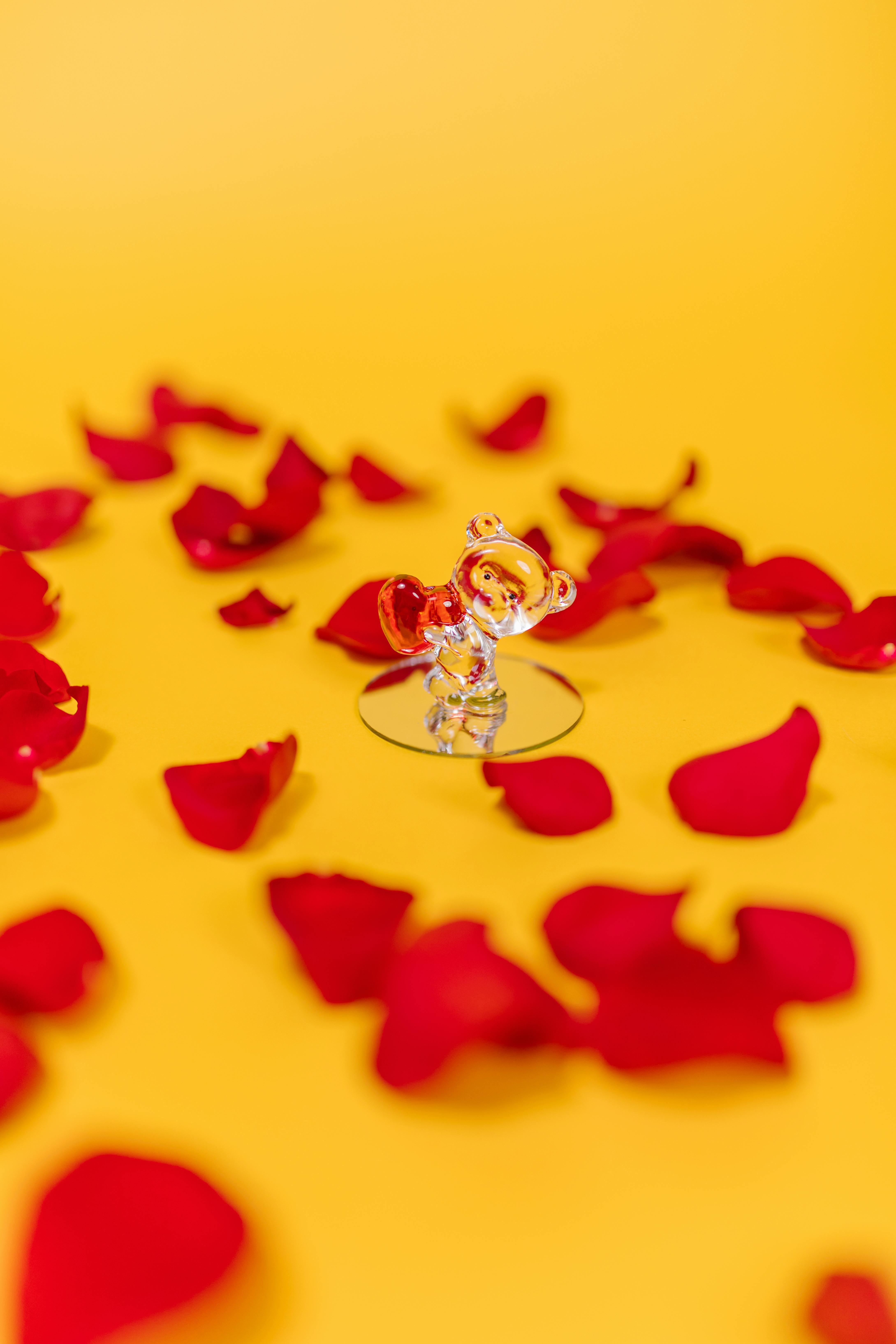 Elegant crystal teddy bear figurine surrounded by red rose petals on a vibrant yellow background, ideal for romantic themes.