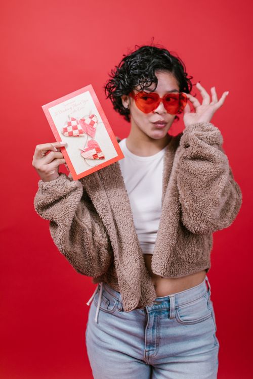 Free Woman in fuzzy jacket holding a Valentine's card and wearing red sunglasses against a red backdrop. Stock Photo